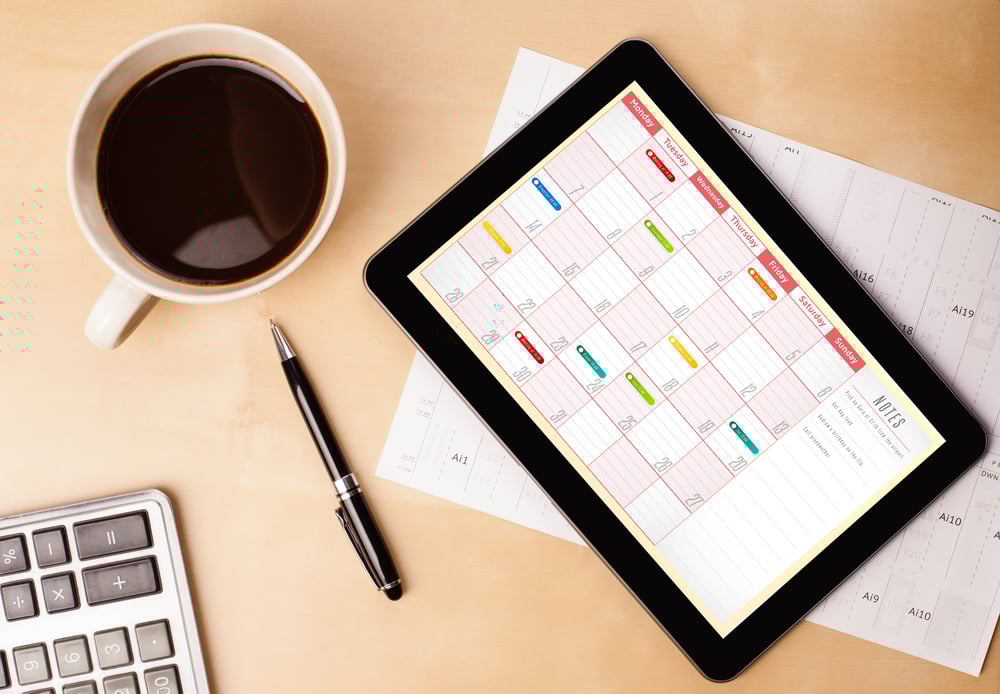 A desk with a pen, cup of coffee, and electronic tablet displaying a weekly calendar.