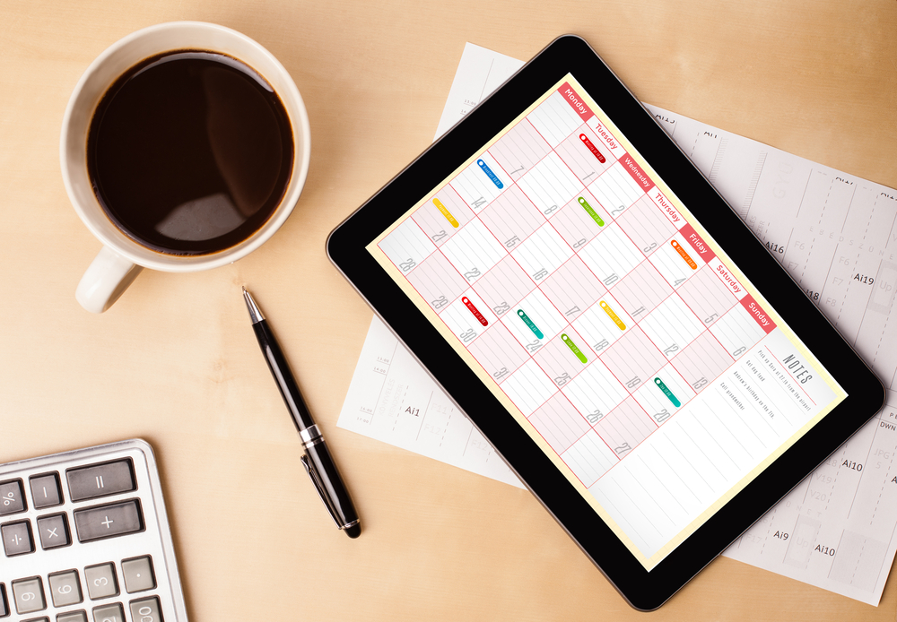 A desk with a pen, cup of coffee, and electronic tablet displaying a weekly calendar.